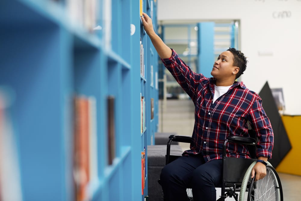 Portrait of disabled student in wheelchair choosing books while studying in college library
